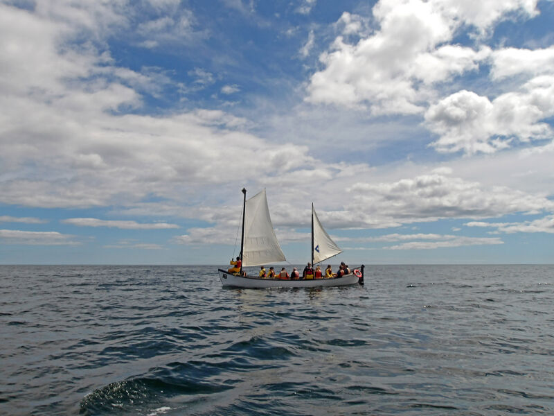 The image shows a sailboat navigating through the ocean under a partly cloudy sky. The boat is carrying a number of passengers. The sea is calm with gentle waves, and the sky features a mix of blue patches and fluffy white clouds. The overall scene suggests a pleasant day for sailing.